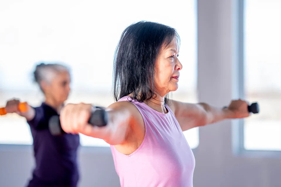A focused, middle-aged Asian woman in a pink tank top performs arm-extension exercises with dumbbells in a bright studio, while another participant exercises in the background, highlighting a group fitness setting.