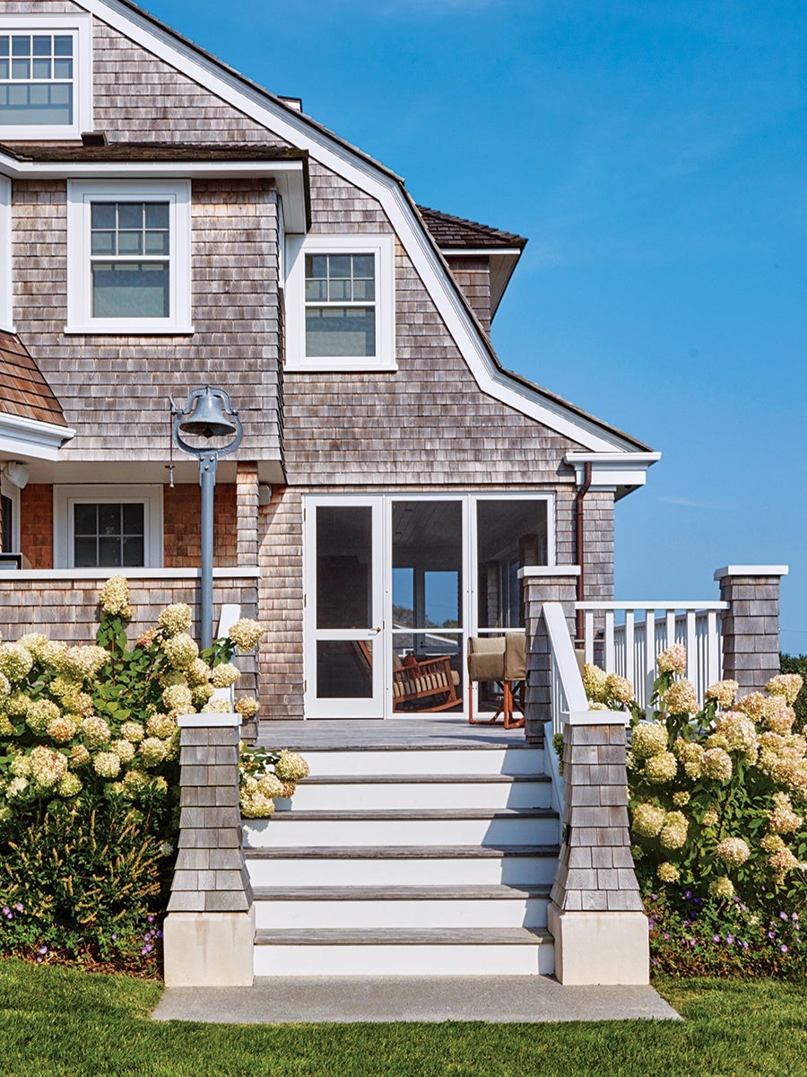 Wooden house exterior with weathered gray shingles, white-framed windows, and a white staircase leading up to a porch. The porch has a glass door and is flanked by lush hydrangea bushes with pale yellow flowers. A metal bell on a post stands to the left of the stairs, and the sky is clear and blue.