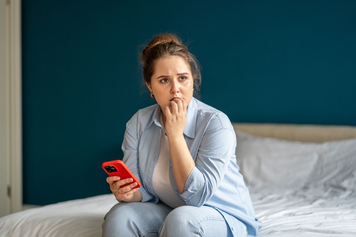 Woman sitting on bed with worried glance, gnawing nails, holding smartphone