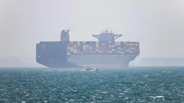 A large cargo ship seen passing through a body of water. The view of the ship is slightly obstructed through fog and a smaller vessel can be seen in front of it