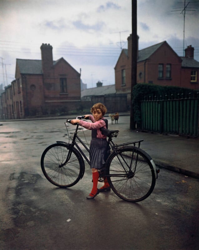 A young girl with short hair, wearing a pink sweater, gray skirt, and red knee-high socks, stands on a wet street holding a black bicycle. Brick houses and a green fence are in the background under a cloudy sky.