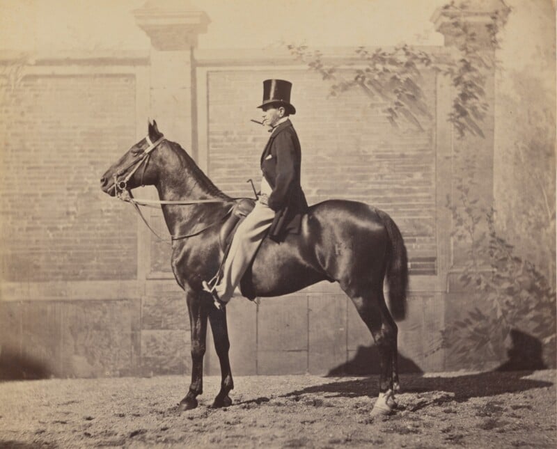A man in formal attire and a top hat sits upright on a dark horse, holding the reins. They are positioned sideways in front of a tall brick wall with decorative stone pillars and some leafy shadows.