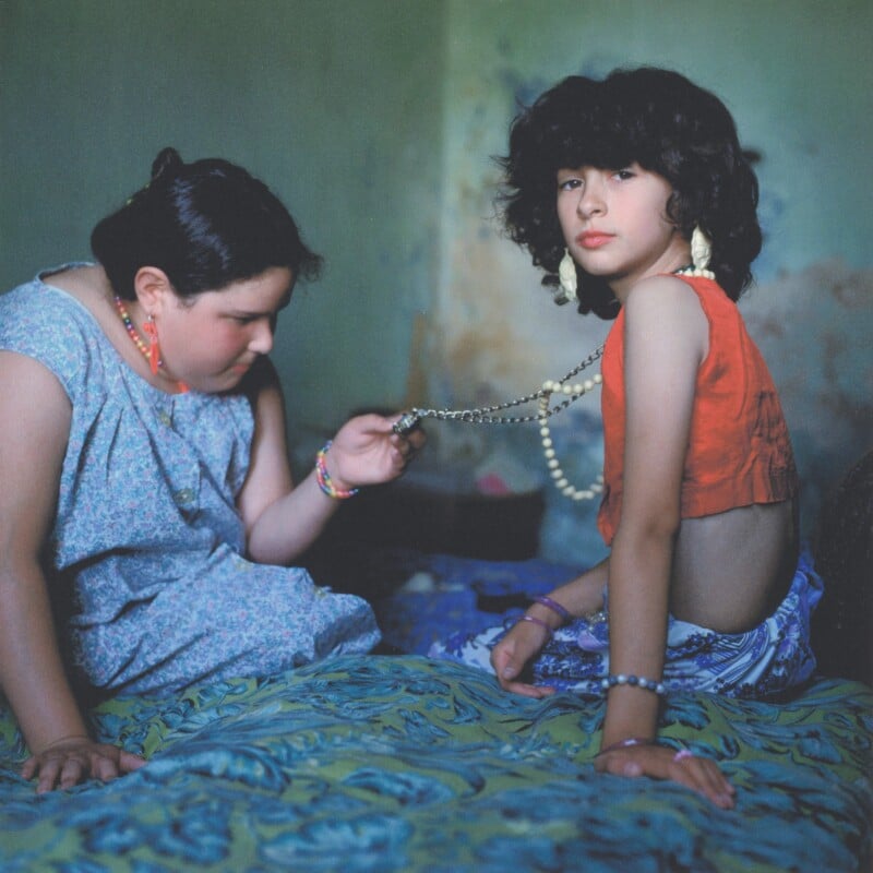 Two children sit on a bed; one wears a blue dress and looks down while holding a beaded necklace that the other, in a red top and patterned skirt, is wearing. The scene is softly lit with a green wall in the background.