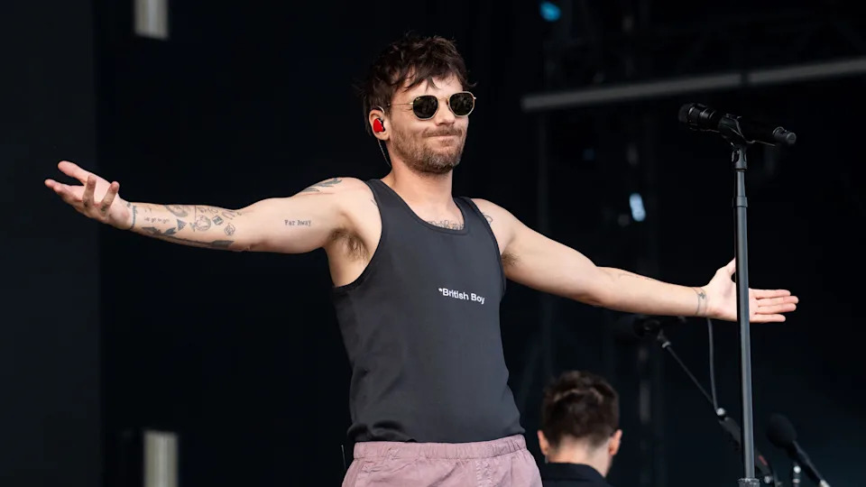 Louis Tomlinson performs on stage wearing a black t-shirt with "British Boy" written on it. He is also wearing sunglasses and pale pink trousers.