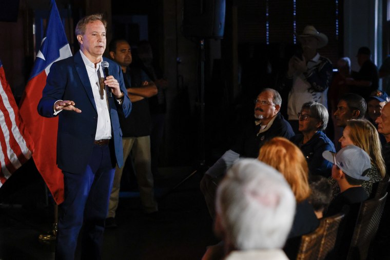 Ken Paxton standing on a stage in a darkened room with a microphone.