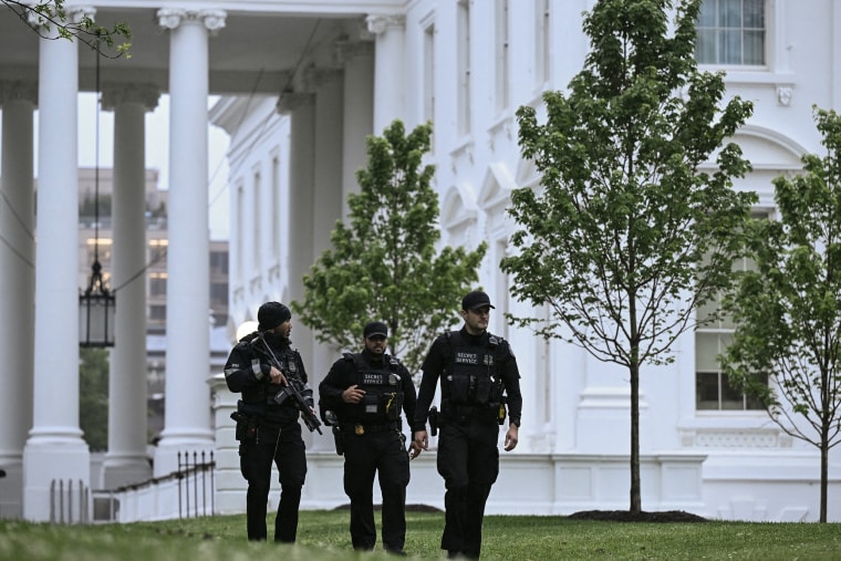 Three secret service agents wearing black uniforms on the White House lawn at dusk.