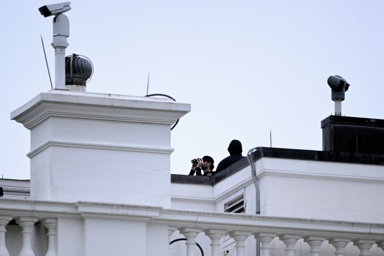 A secret service agent with binoculars on the roof of a building at the White House.
