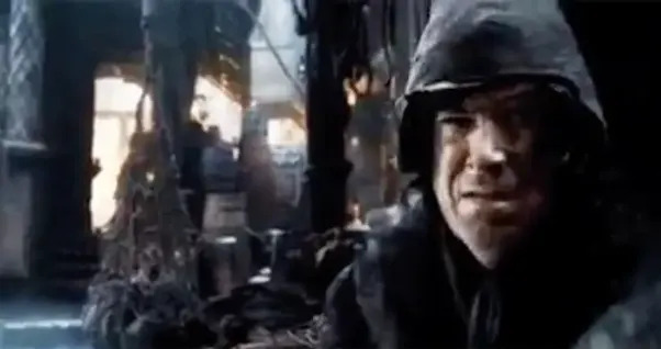 Scene from a dark fantasy film showing a hooded character with a tense expression, surrounded by mysterious and shadowy props