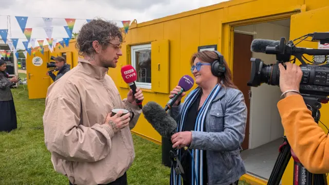 Tom Grennan is interviewed at the parade in front of a yellow porta cabin. He is wearing a beige waterproof jacket and brown sunglasses.
