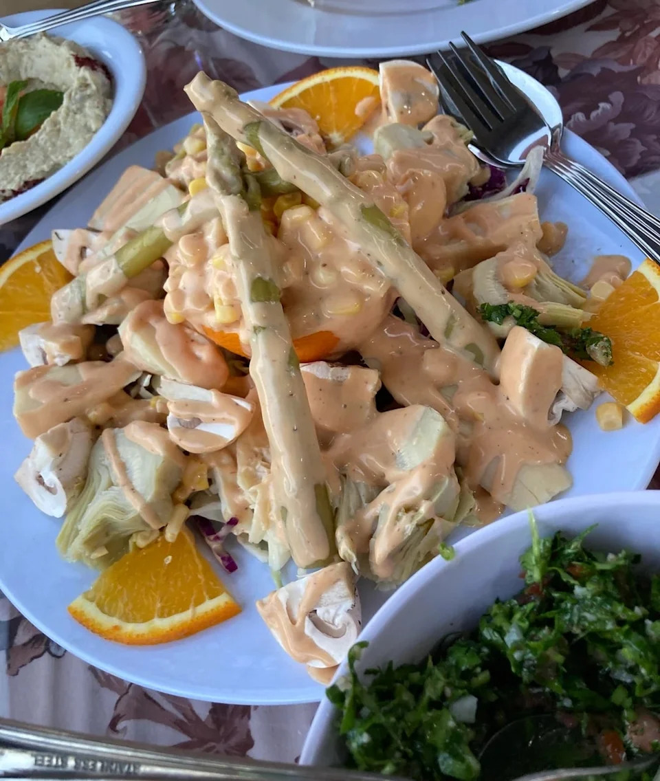 Plate of salad with asparagus, artichokes, orange slices, and dressing. Accompanied by hummus and tabbouleh in small bowls
