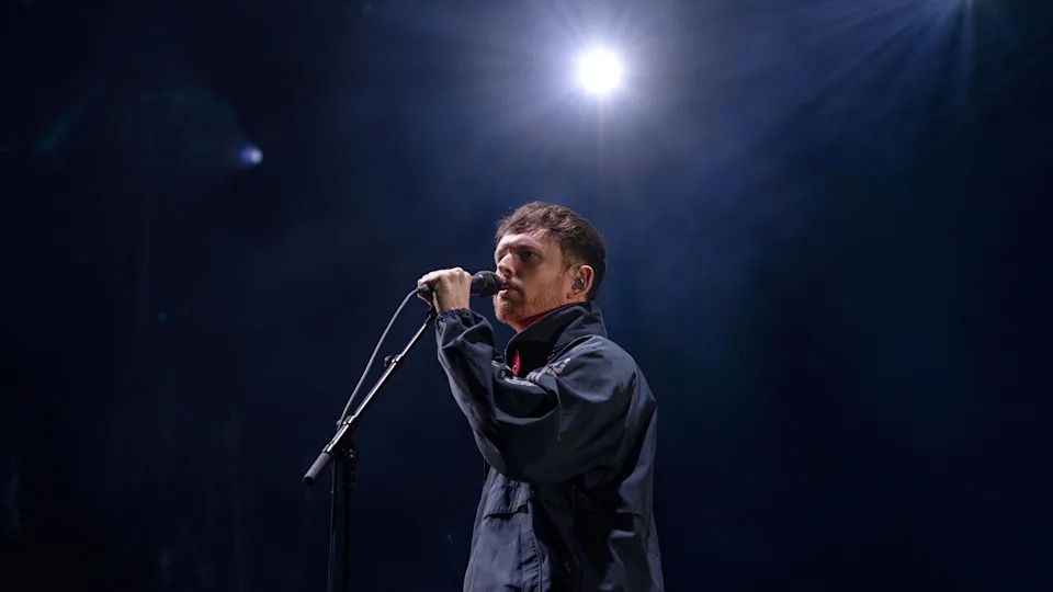 James Blake sings into a microphone on stage infront of a dark background. He is wearing a dark sports jacket.