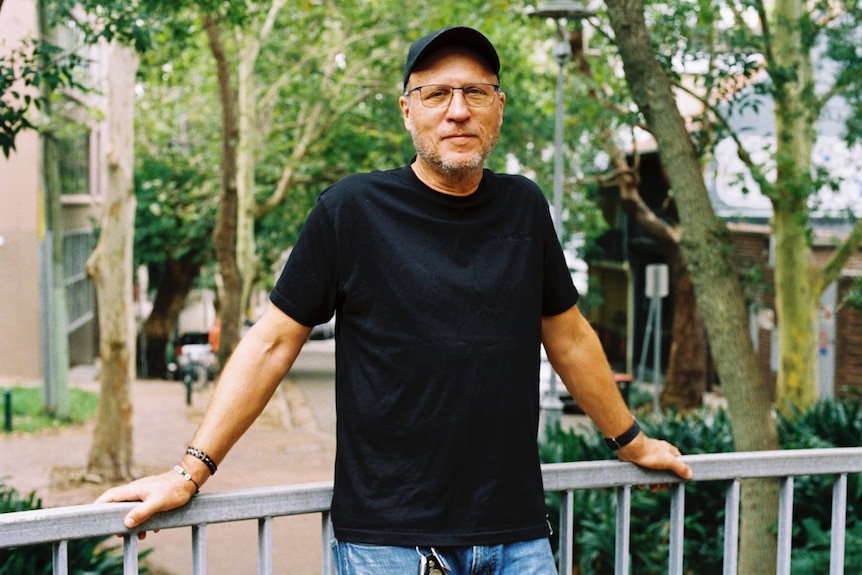 A man wearing a T-shirt, cap and jeans leans again a metal railing in a leafy suburb.