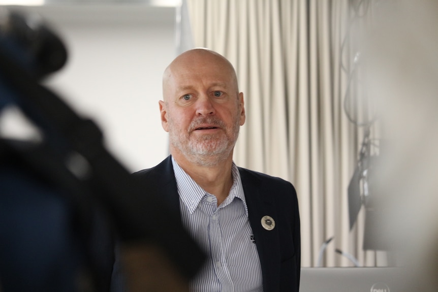 Doug wears a shirt and suit jacket and watches others.