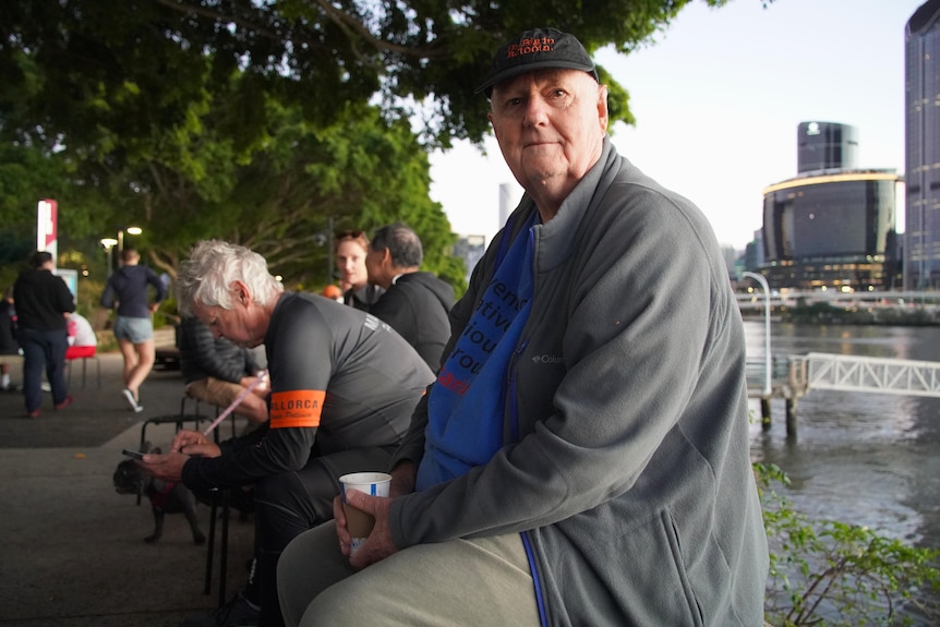 A man sits on a wall next to the Brisbane River holding a cup of coffee.