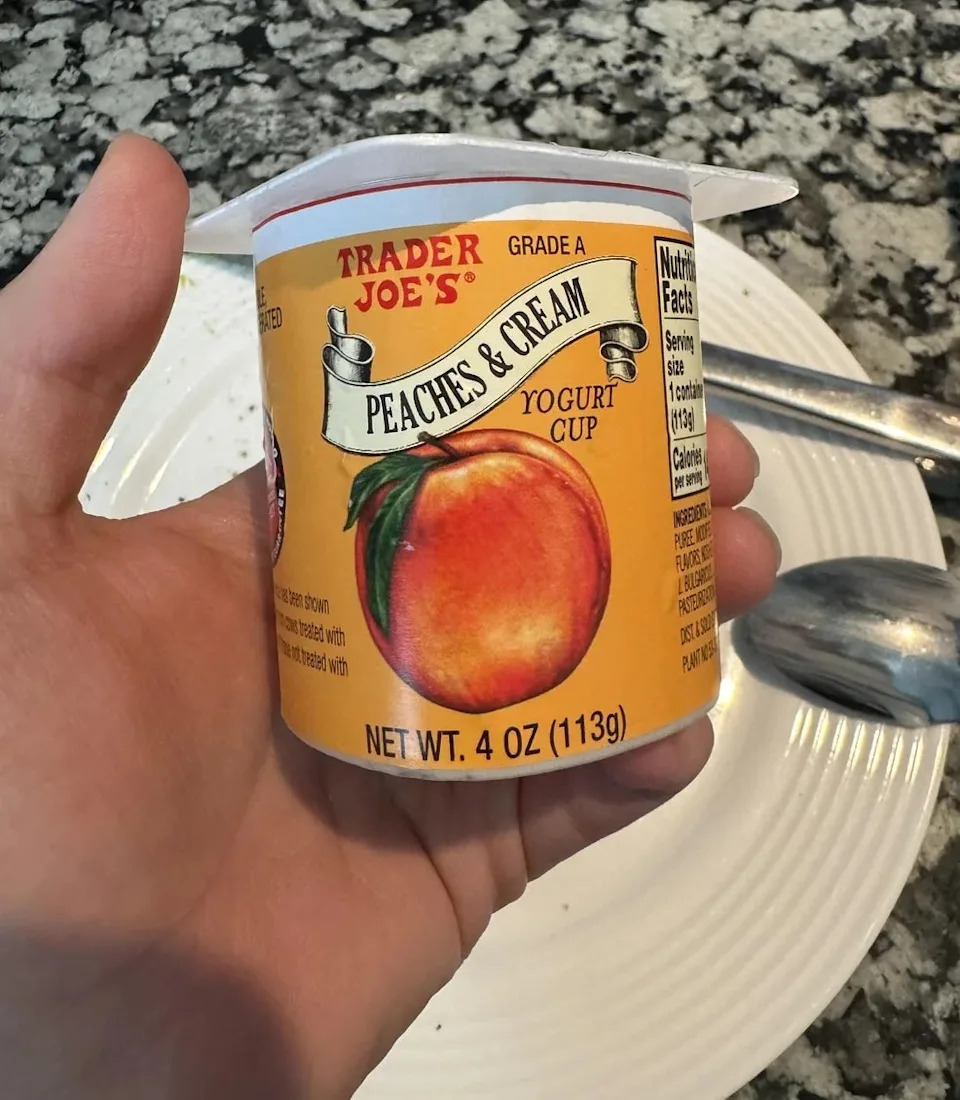 Person holding a Trader Joe's Peaches & Cream yogurt cup over a kitchen counter with a spoon nearby