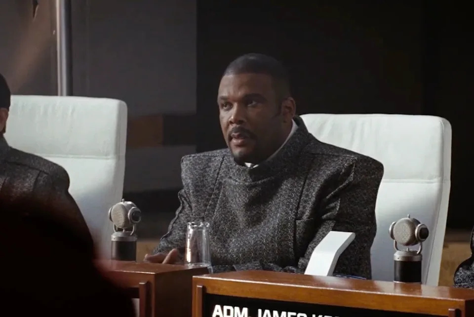 A person in a futuristic setting speaks at a conference table, wearing a sleek modern suit, suggesting a sci-fi or action film scene