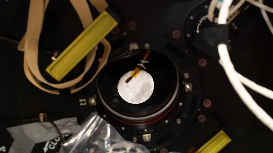 A view of a circular, gray moon through a round window in a space capsule.