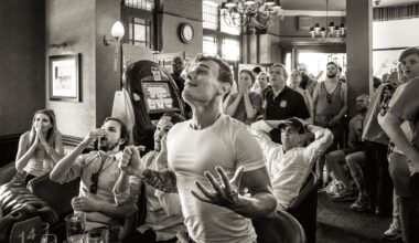 Black and white image of a crowded pub with people reacting emotionally, some seated and others standing near a slot machine