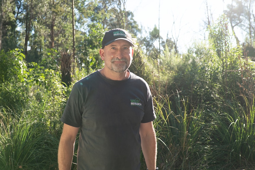 A man with a beard, wears a black shirt and cap, standing in the forest. 