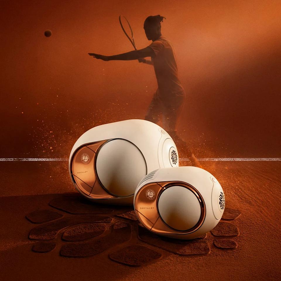 Two white and copper Devialet Phantom speakers on a clay tennis court with a tennis player silhouette in the background.