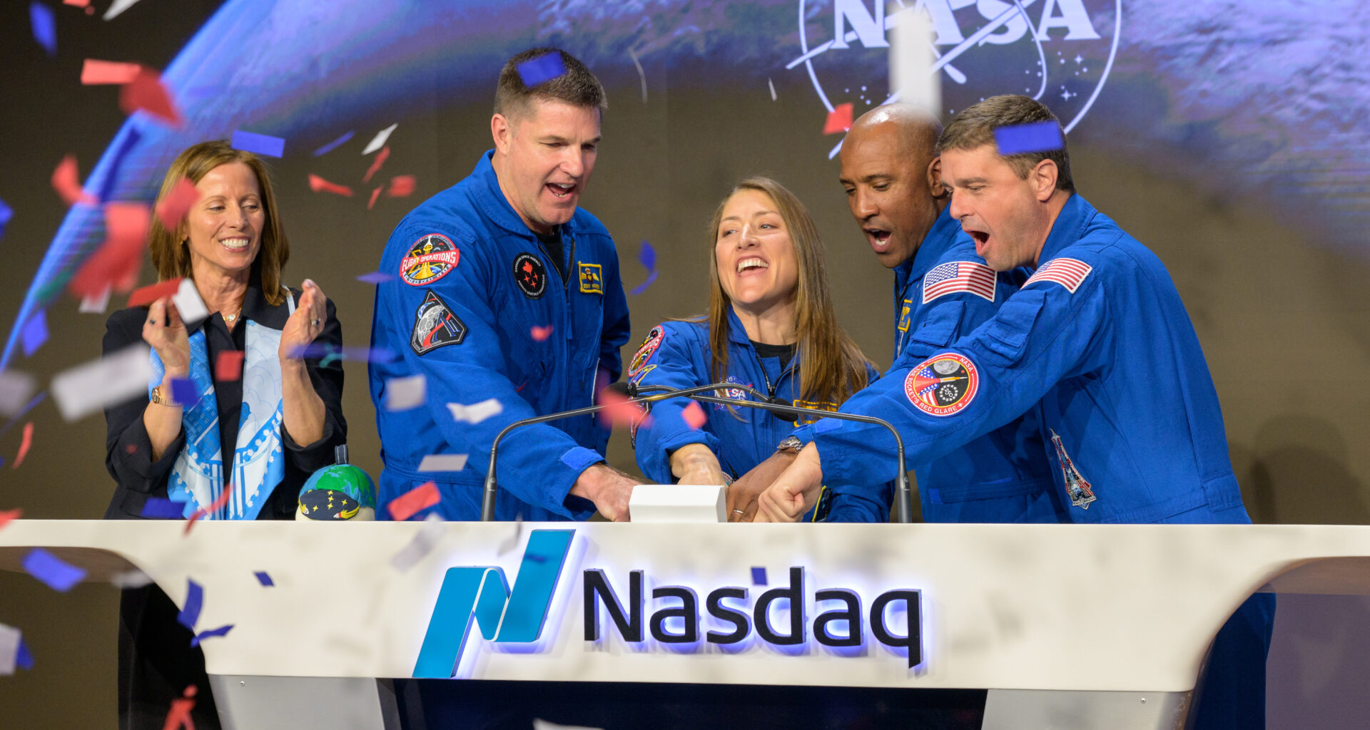 Nasdaq Chair and Chief Executive Officer Adena T. Friedman, left, and NASA’s Artemis II crewmembers CSA (Canadian Space Agency) astronaut Jeremy Hansen, and NASA astronauts Christina Koch, Victor Glover, and Reid Wiseman, right, ring the closing bell of the Nasdaq market session. They're standing behind a wide white podium with the Nasdaq logo and the word "Nasdaq." There is a screen on the front of the podium displaying the NASA meatball logo. Friedman is standing a little further away from the podium. Red, white, and blue confetti falls from the air on the left side of the image.