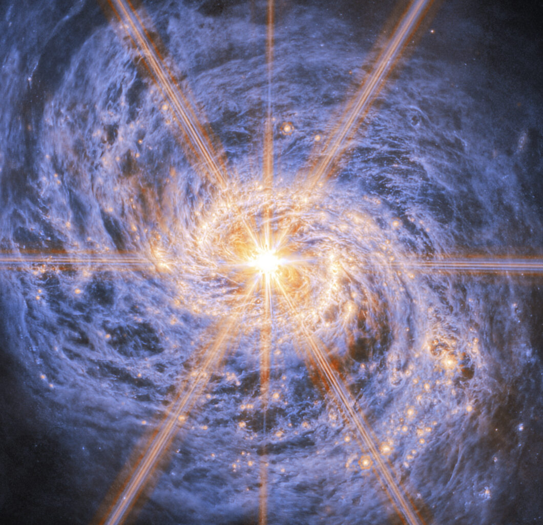 A spiral galaxy shown in mid-infrared light. The image is dominated by an extremely bright glow from the galaxy’s nucleus. Six large and two smaller rays of light emit from the center, which are diffraction spikes created by the telescope’s optics. The galaxy’s spiral arms are visible by two lines of glowing orange bubbles which whirl out into the disc. Swirling blue clouds of dust make up the rest of the galaxy.