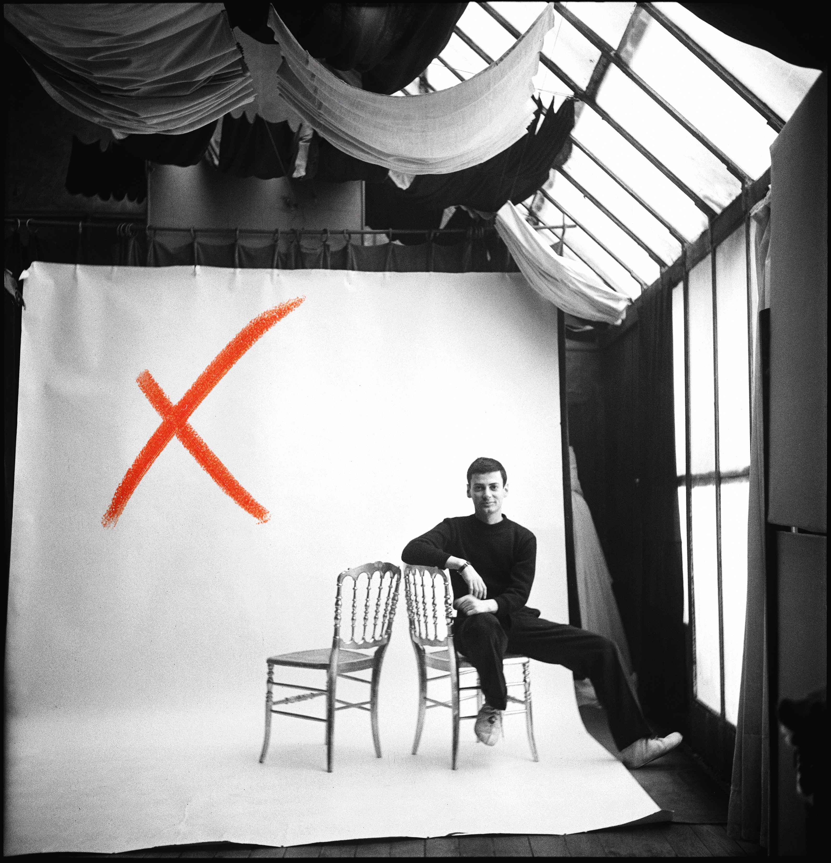 A young Richard Avedon in black sits casually across two ornate chairs before a white studio backdrop marked with a large red painted X.