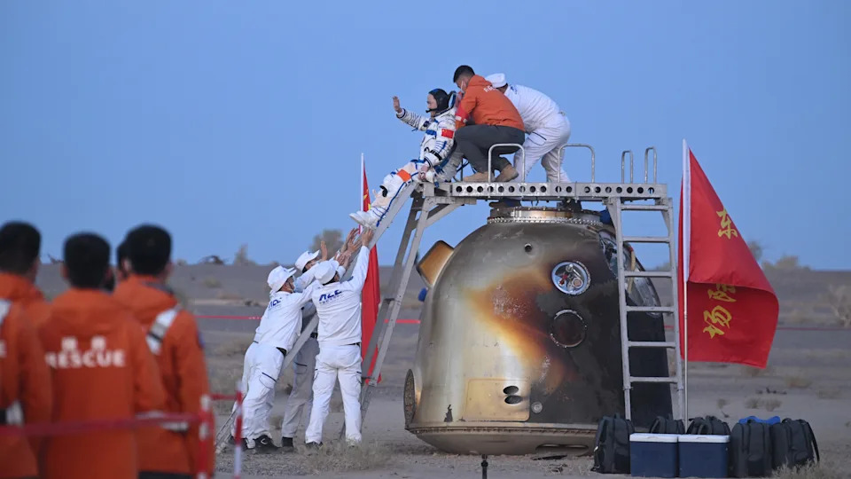 three people wearing help an astronaut wearing a white spacesuit down from the top of a golden spacecraft in a desert landscape