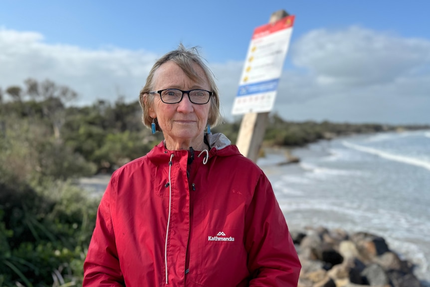 A woman with glasses wearing a red jacket standing near a beach