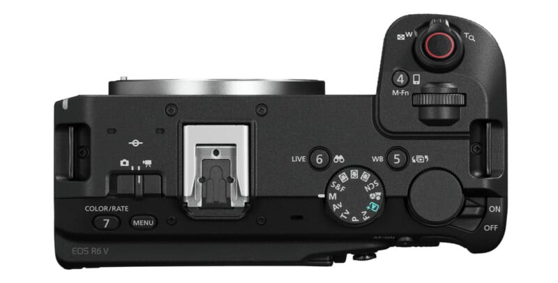 Top view of a black digital camera showing buttons, dials, an LCD panel, hot shoe mount, and a mode dial with various settings. The camera lens is not attached.