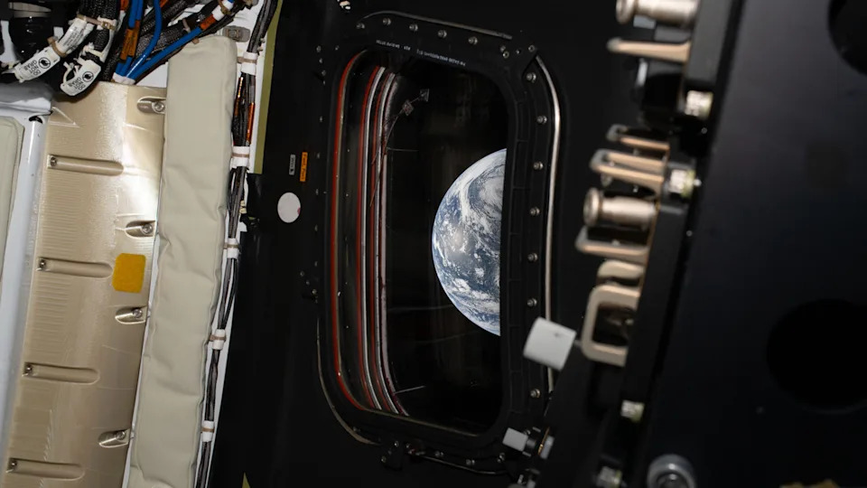 A view of Earth, its blue and white colors, are seen through a rectangular view port window inside a space capsule.