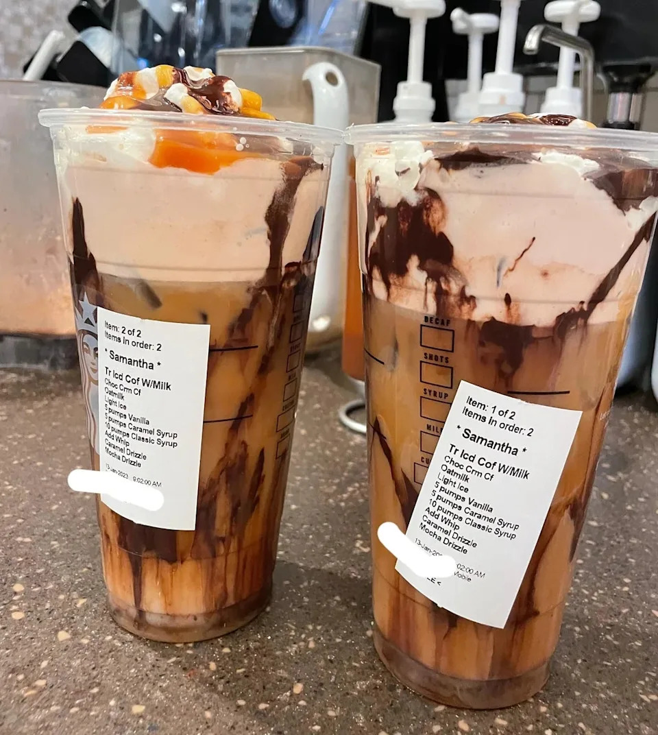 Two iced coffee drinks topped with whipped cream, caramel drizzle, and chocolate syrup, labeled with customization details on the cups