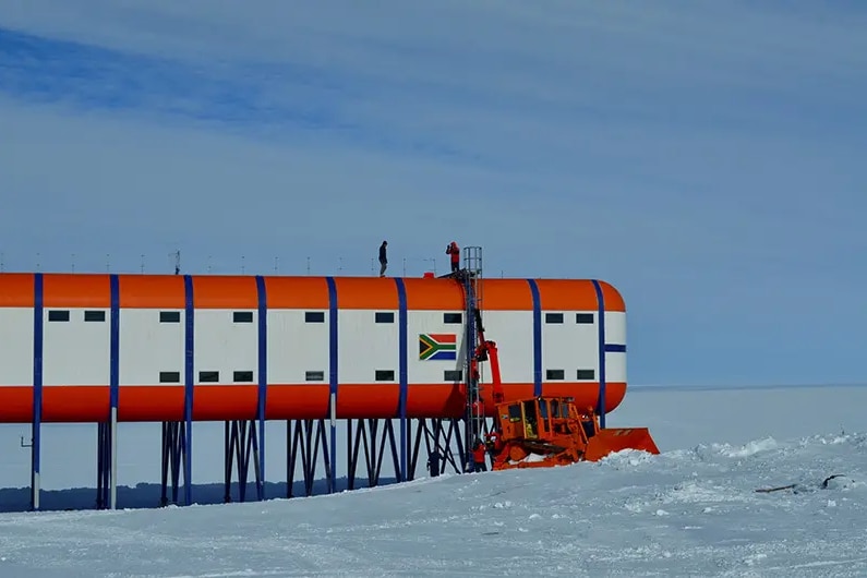 A side view of an orange and white building in a snowy, Antarctic landscape. There is a South African flag on the building.