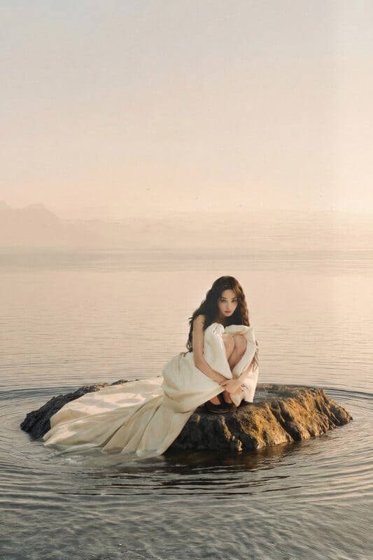 A woman in a long, flowing white dress sits curled up on a small rock surrounded by calm water, with a hazy sky and distant mountains in the background.