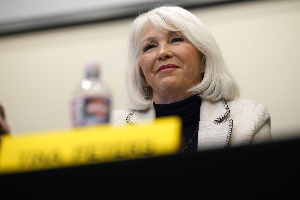 Tina Peters, who has white hair, is seated, wearing a white jacket and black top, with a water bottle and a yellow name tag in front of her.