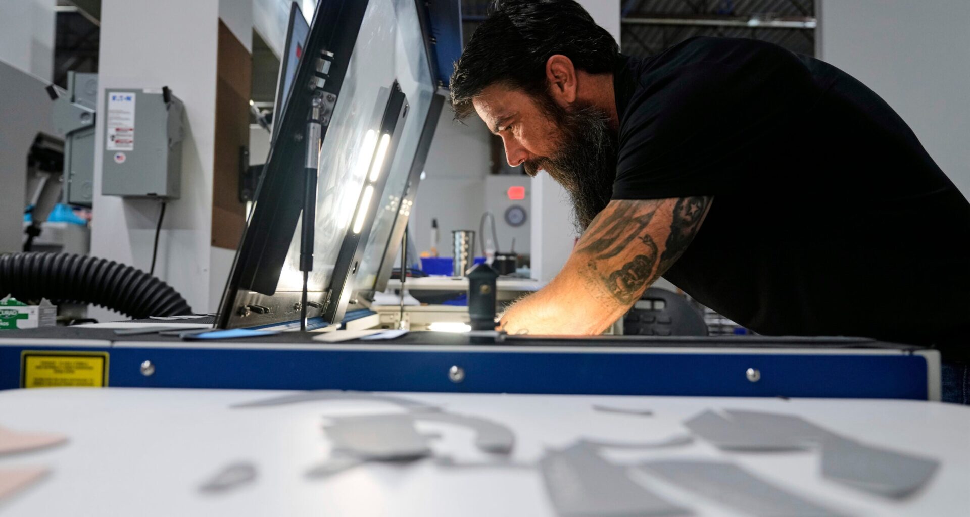 A man with a beard and tattoos operates machinery under a light in an industrial workshop.