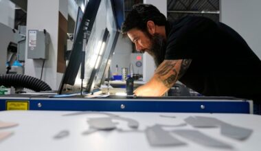 A man with a beard and tattoos operates machinery under a light in an industrial workshop.