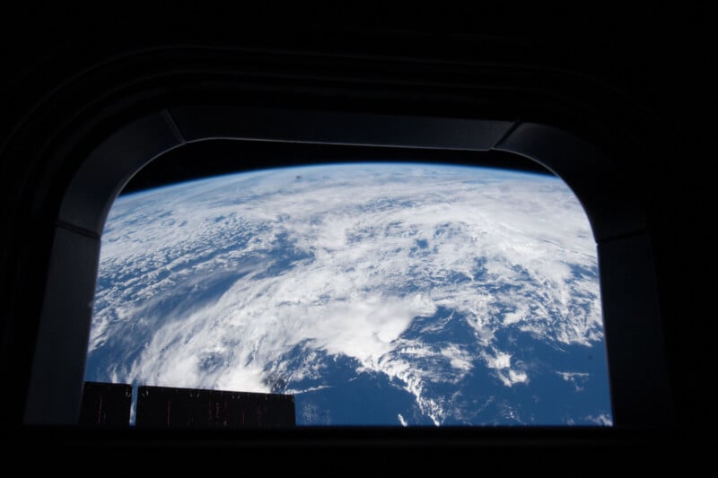 View of Earth’s cloudy atmosphere and blue surface seen through a window from space, with part of a dark spacecraft structure visible around the edges of the frame.