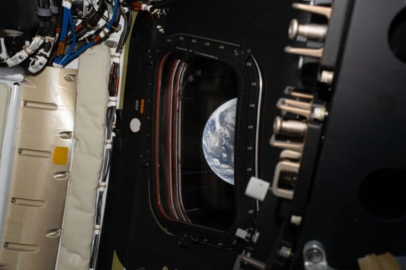 View of Earth through a window from inside a spacecraft, surrounded by panels, cables, and machinery parts. The planet appears partially visible against the blackness of space.