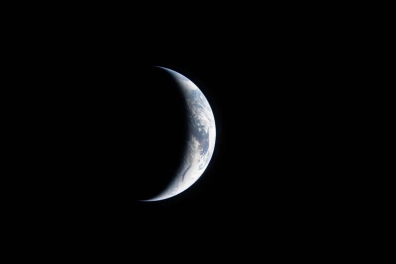 A crescent-shaped Earth is partially illuminated against a black background, with clouds and landforms visible on the bright edge, resembling a crescent moon.