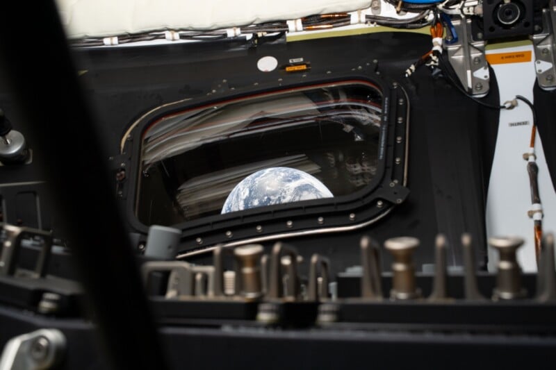 View of Earth through a window from inside a spacecraft, with various buttons, switches, and equipment panels visible in the foreground.