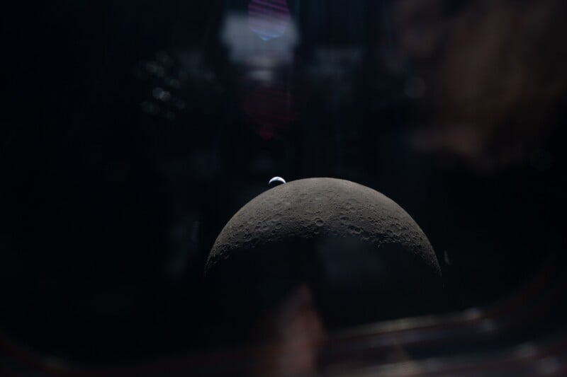 A view of the Moon's dark, cratered surface seen from space, with Earth appearing as a small blue and white sphere above the Moon’s horizon against a black background.