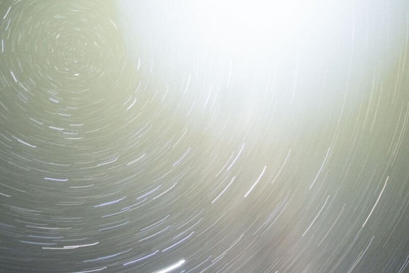 Long-exposure photograph of the night sky showing circular star trails, creating a spiral pattern, with a bright, diffused light source at the upper right corner. The background is a soft gradient from light to dark.