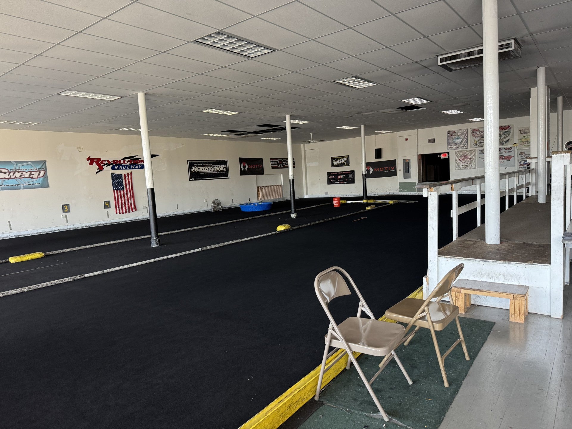 Indoor RC car track with black racing surface, yellow lane dividers, wall banners, two folding chairs, and a small ramp leading to an elevated walkway.