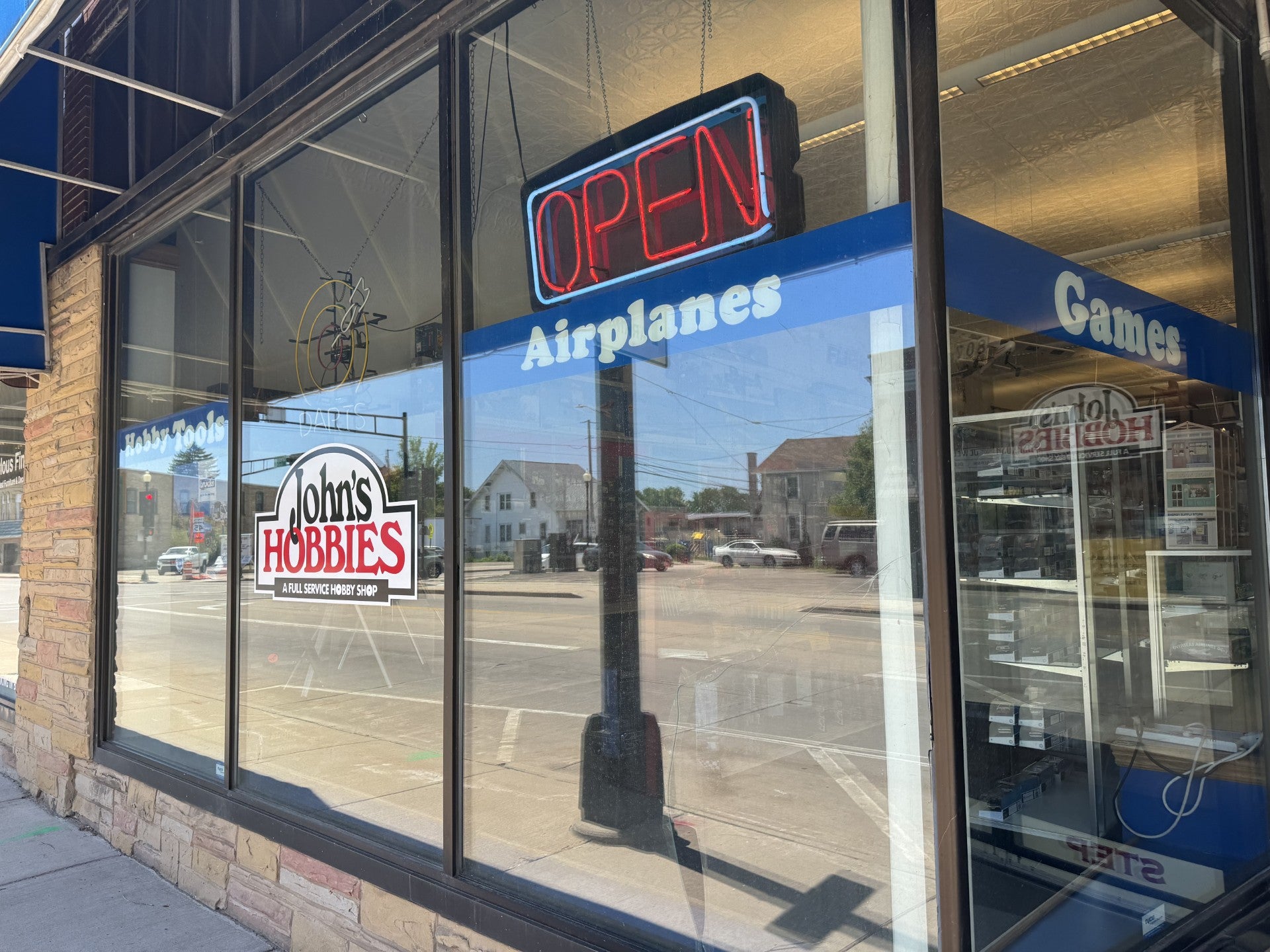 Storefront window of Johns Hobbies with a neon OPEN sign; window text includes Airplanes and Games; street and buildings reflected in the glass.