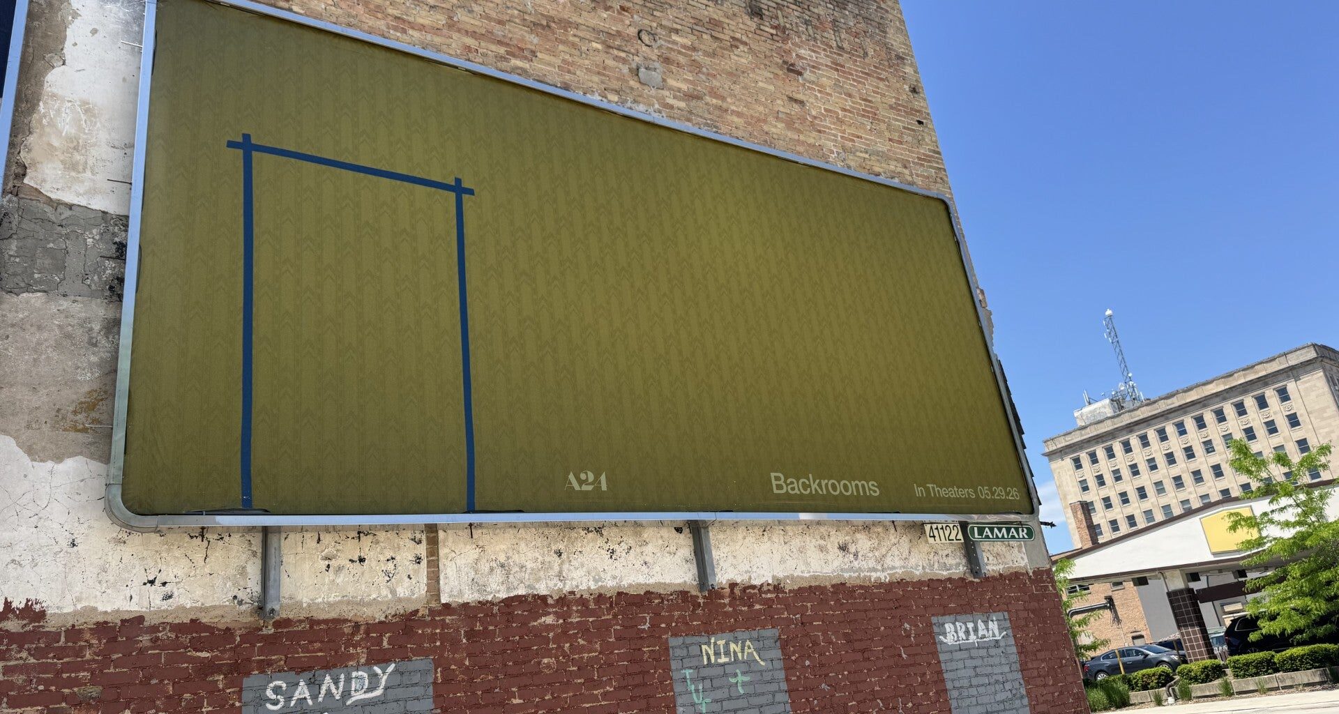 A yellow-green billboard with a faint Backrooms pattern and blue tape outline is mounted on a brick wall in an urban setting under clear skies.