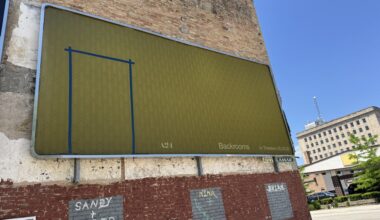 A yellow-green billboard with a faint Backrooms pattern and blue tape outline is mounted on a brick wall in an urban setting under clear skies.
