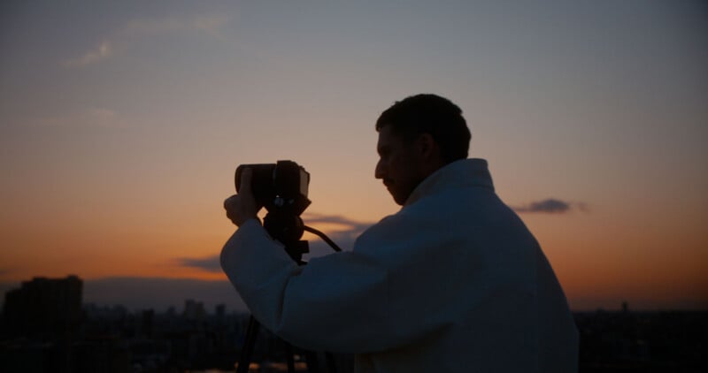 A person in a light jacket adjusts a camera on a tripod, silhouetted against a colorful sunset sky with city buildings in the background.