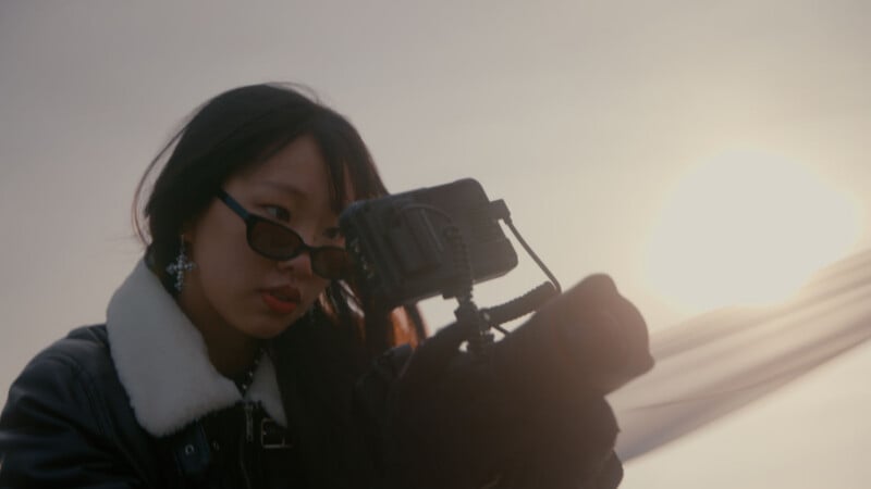 A woman wearing sunglasses and a black jacket with a white collar looks closely at a camera, with the sun setting in the background and a soft, hazy sky.