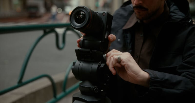 A person adjusts a digital camera mounted on a tripod outdoors, wearing a dark jacket and a ring on their finger. The background is blurred, showing a street and railing.